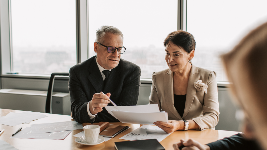Two executives at a boardroom table, reviewing printed reports and charts together